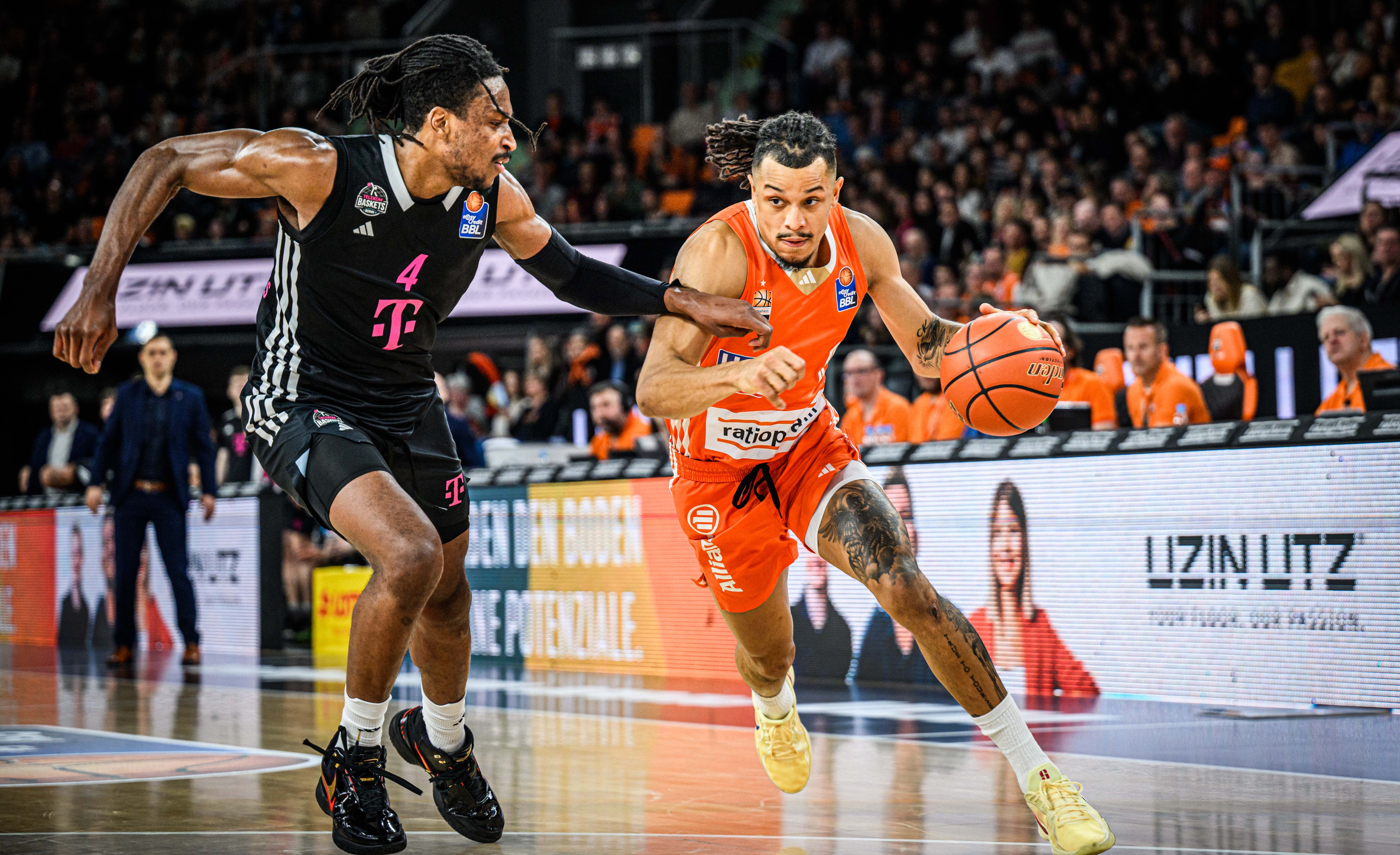 Basketballspieler im orangefarbenen Trikot im Angriff, dynamische Spielsituation vor Uzin Utz Bandenwerbung